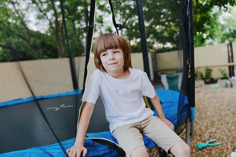 Playful boy sitting on a trampoline, enjoying a sunny day outdoors with a smile, surrounded by greenery and a fun atmosphere.