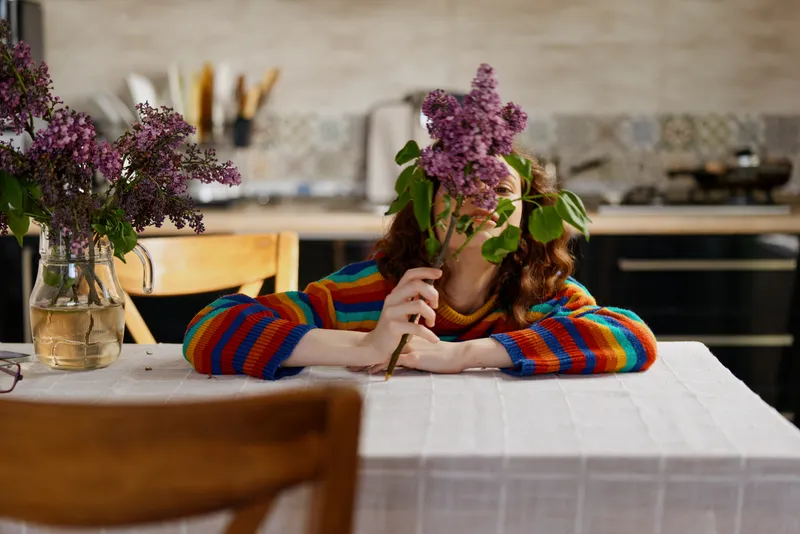 A woman sits at a dining table in a cozy home, bouquet held over her face as she rests her chin on her arm. The bright kitchen in the background adds warmth and a moment of introspection.