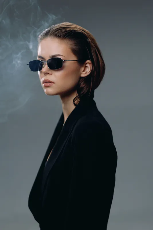 Stylish woman with short hair wearing dark sunglasses and black jacket against grey background, emitting mysterious vibe.