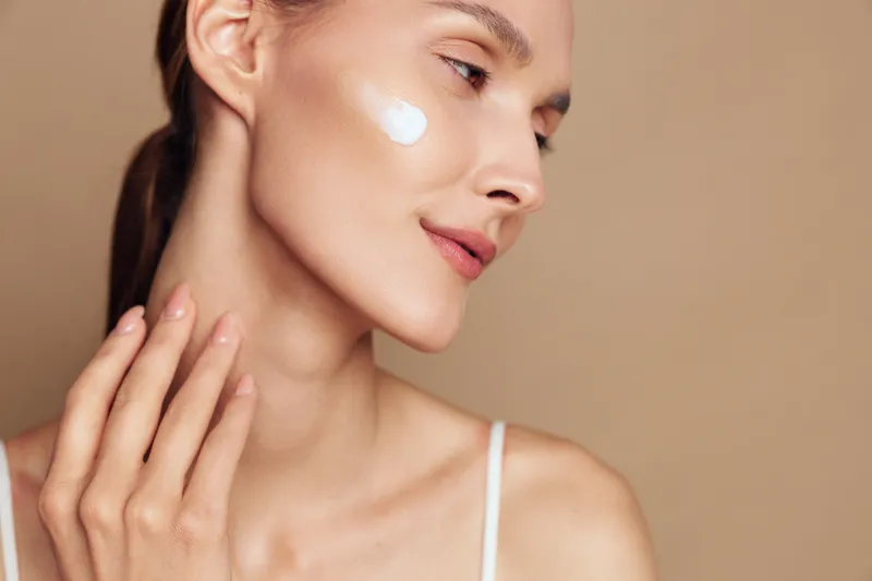 A young woman applies moisturizing cream to her cheek with her hand, showing smooth skin and a calm expression against a neutral beige background, concept for skincare and beauty.