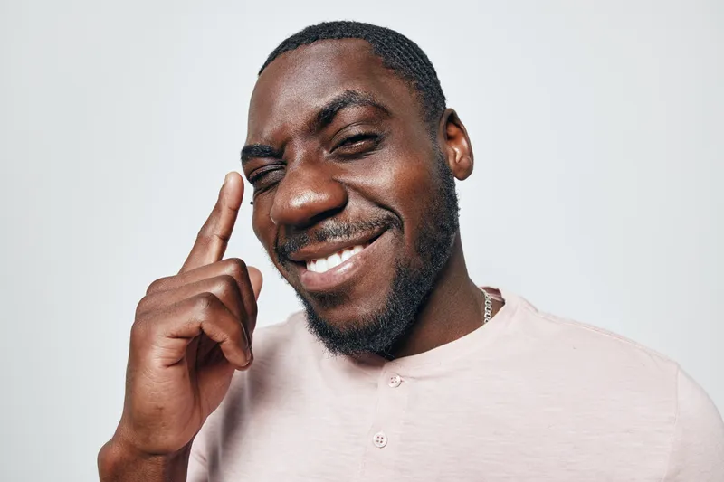 African American man with dark skin smiling and pointing finger to head in casual shirt on white background. Portrait of confident black male with short hair and beard.
