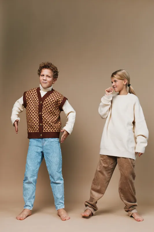 Two children pose in cozy vintage outfits against a neutral backdrop, highlighting a knitted vest, oversized sweater, denim jeans, and corduroy pants in soft earth tones.
