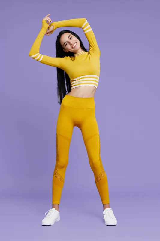 Sport fitness woman stretching in bright yellow activewear, smiling and posing against solid purple background, promoting healthy lifestyle and flexibility.