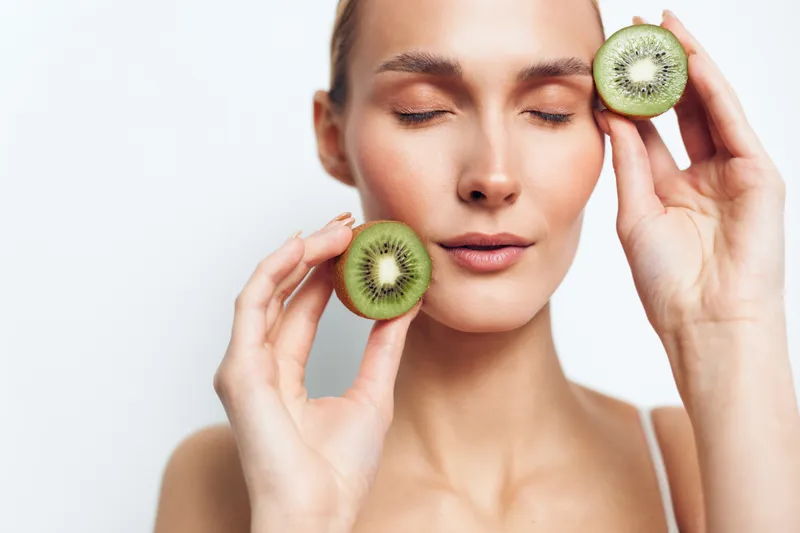 Woman kiwi skincare beauty spa facial wellness fruit slices near cheeks with eyes closed on clean white background soft natural light calm healthy studio portrait