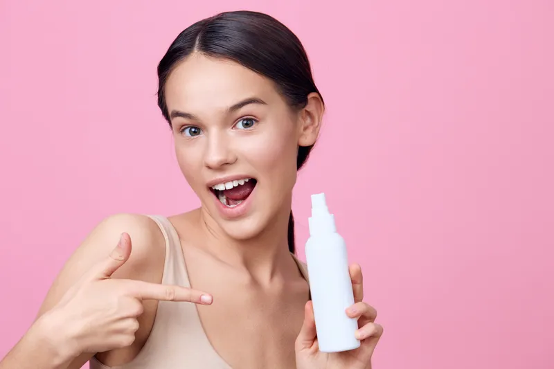 Smiling young woman with long hair showcasing a skincare product against a pink background, emphasizing beauty and confidence in a health focused concept