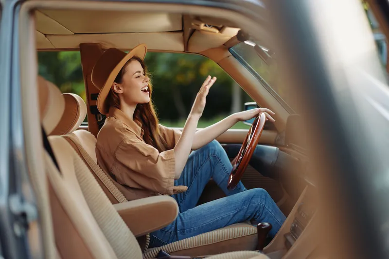 Young woman driving a vintage car, wearing a stylish hat and casual clothes, expressing joy with a smile and waving her hand, perfect for travel themes.