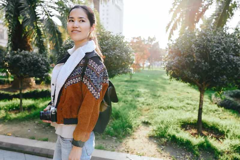 Woman with camera in a sunny park, wearing a color block jacket and scarf, backpack on shoulder, as autumn light makes trees glow during a casual outdoor outing