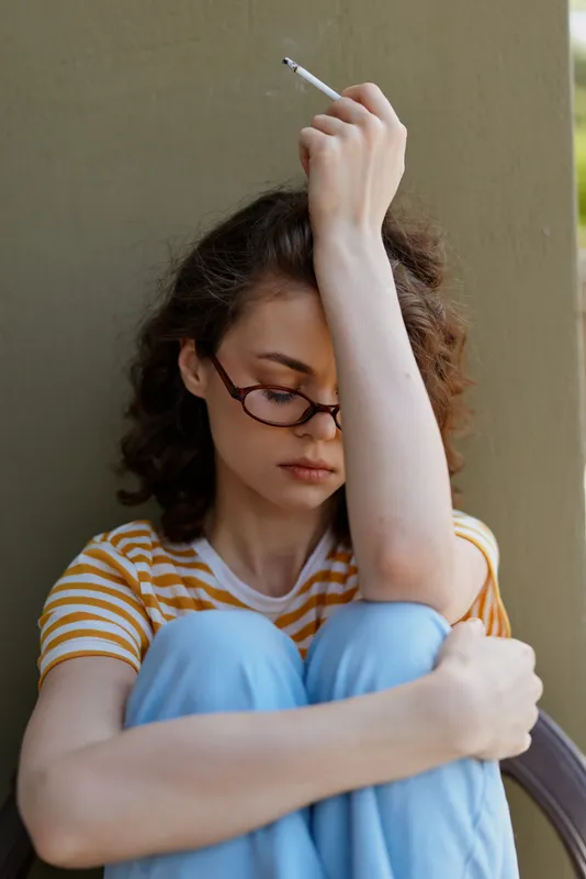 Young girl with glasses sits against a wall, wearing a striped shirt and blue jeans, feeling sad and lonely, holding a cigarette above her head, reflecting a pensive mood.