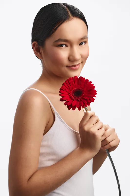 asian woman holds a bright red flower close to her face in a studio portrait, wearing a light top, soft lighting, clean backdrop, creating a calm, elegant and uplifting atmosphere.