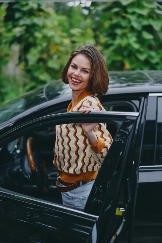 Joyful woman steps from a sleek car, smiling at the camera, wearing a patterned knit sweater and jeans in a vibrant outdoor setting.