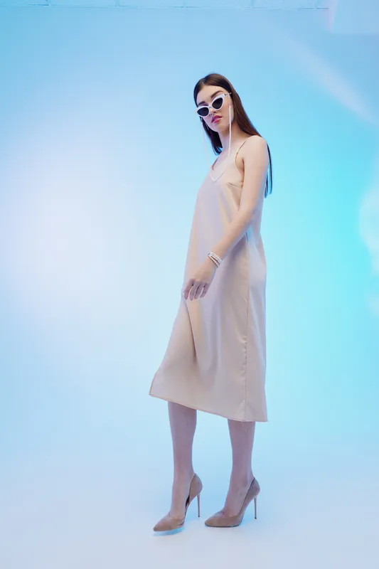 Young woman in a beige dress and sunglasses poses confidently in a studio with blue lighting. Concept of beauty, fashion, elegance, and modern style.