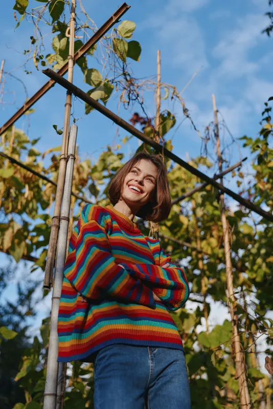 Woman smiling in rainbow sweater in garden outdoors, portrait of happy young woman with crossed arms, casual jeans and short hair near trellis under blue sky.