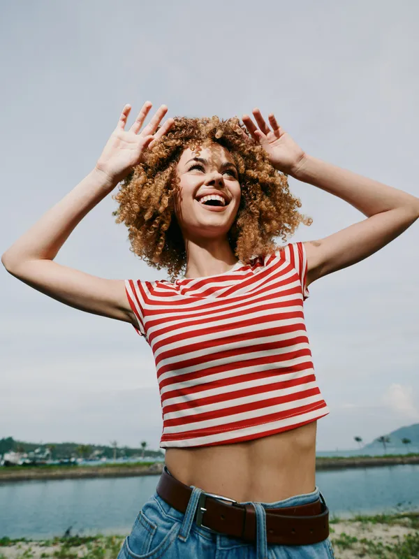 Joyful young woman with curly hair in a red striped tee raises arms in the air, smiling under a bright sky, portraying freedom, energy, and outdoor summer fun.