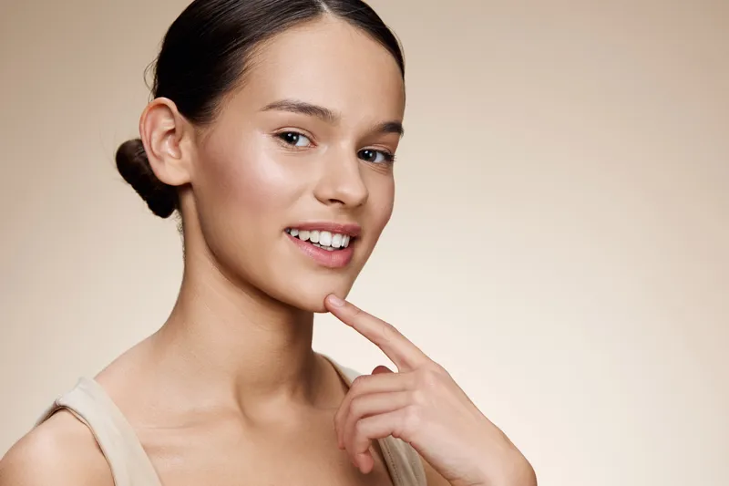 Young woman with a radiant smile touching her chin, showcasing healthy skin in a soft beige background, ideal for beauty and wellness concepts