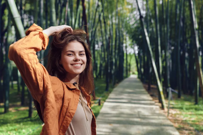 Woman smile portrait on bamboo pathway in park, young traveler wearing orange jacket and backpack, casual outdoor portrait with natural light, happy student exploring nature.