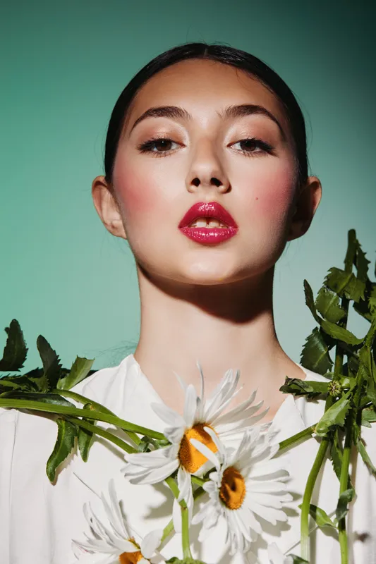 Portrait of a young woman with creative makeup and glossy red lips wearing daisies and greenery, standing against a pastel green background. Fashion and beauty concept.