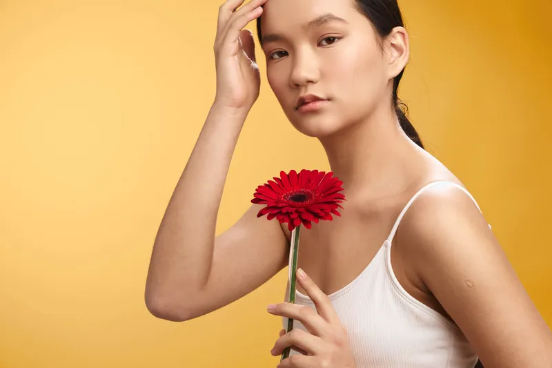 asian woman wearing a white top holds a red flower while posing against a warm yellow backdrop, creating a calm, confident portrait with a gentle, graceful aura.