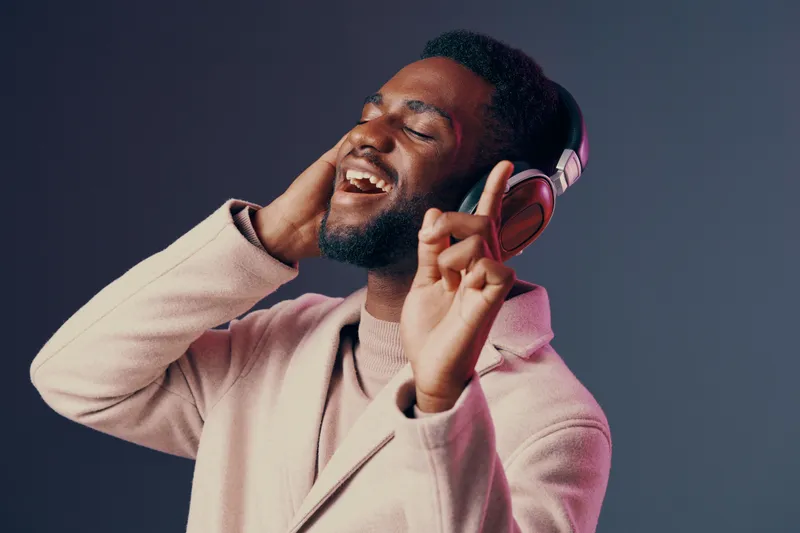 Young Black man enjoying music with headphones, wearing a stylish pink coat on a gradient background, expressing happiness and joy through music