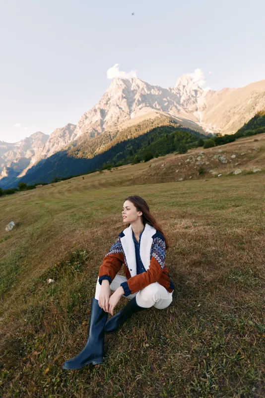 Woman sitting on grassy meadow in front of mountain landscape, wearing warm jacket and boots. Outdoor nature travel scene with peaceful gaze, hiking style and serene autumn light.