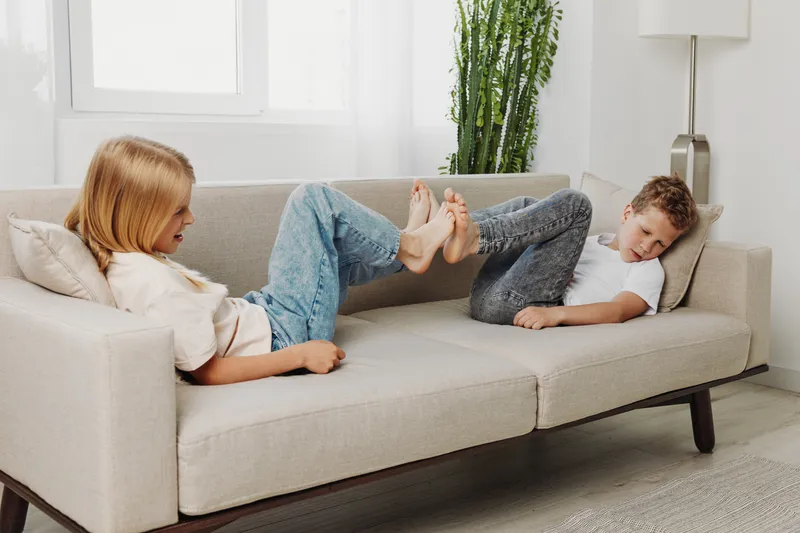 Two children having fun on a beige sofa in a bright living room, playfully touching feet and laughing. Concept of childhood, family bonding, and indoor leisure time.