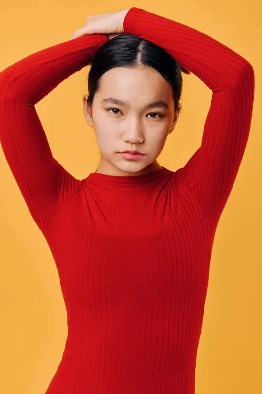 Young woman with black hair wearing red sweater poses standing with arms raised above head against yellow background, looking confident and serious with minimal makeup.