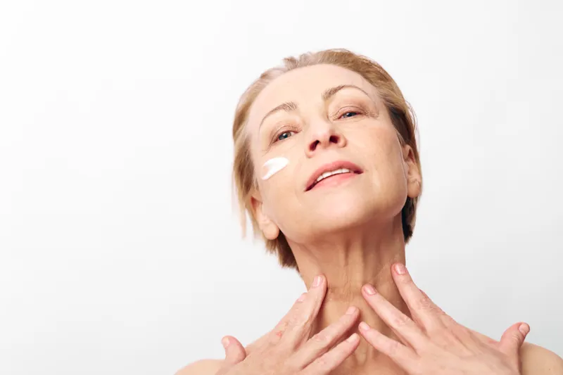 Mature woman applies face cream with fingers on clean skin, looking upwards with gentle smile in bright studio. Concept for skincare, antiaging, selfcare, and natural beauty routine.