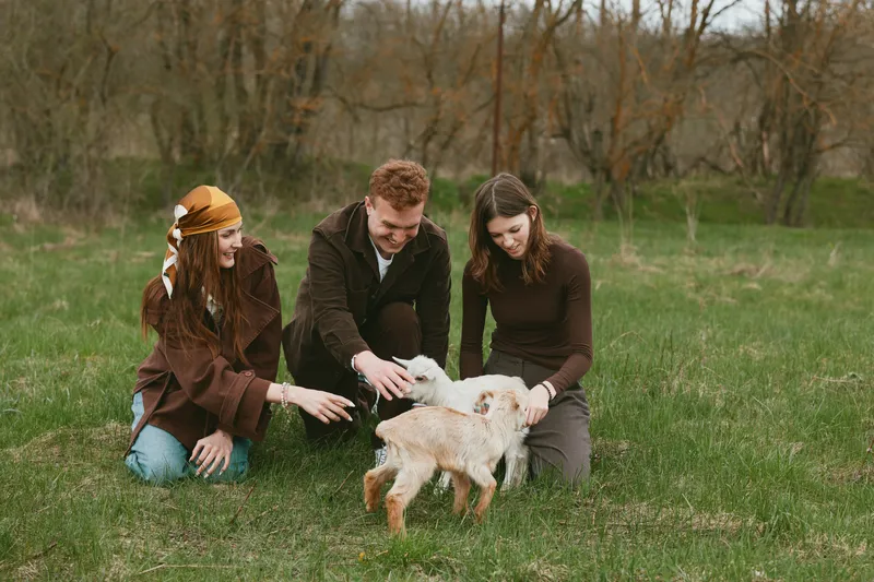 Three young adults enjoy a spring day outdoors playing with two baby goats on green grass in a natural field setting with bare trees in the background.