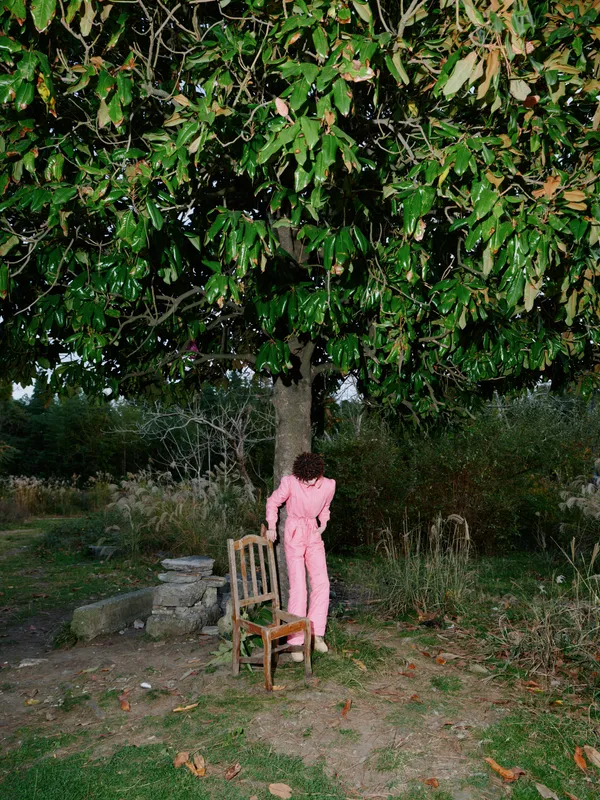 Outdoor scene with a solitary figure in pink standing near a wooden chair beneath a lush tree, a quiet garden setting, dusk light, calm atmosphere and rustic backyard feel.