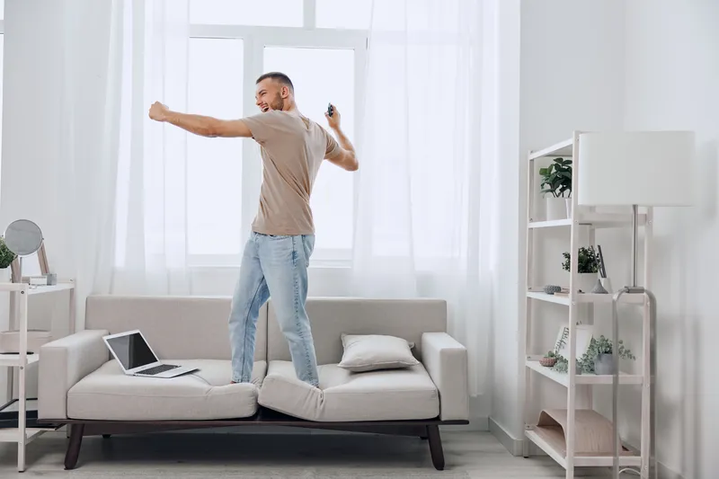 Happy man dancing on couch, enjoying a break from technology, embodying soft masculinity and the freedom of solo living in a modern, airy space.