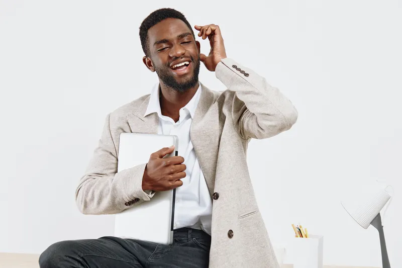 Smiling man holding a tablet and talking on mobile phone, dressed in smart casual attire, sitting in bright modern office environment, conveying communication, technology, business, happiness, and suc