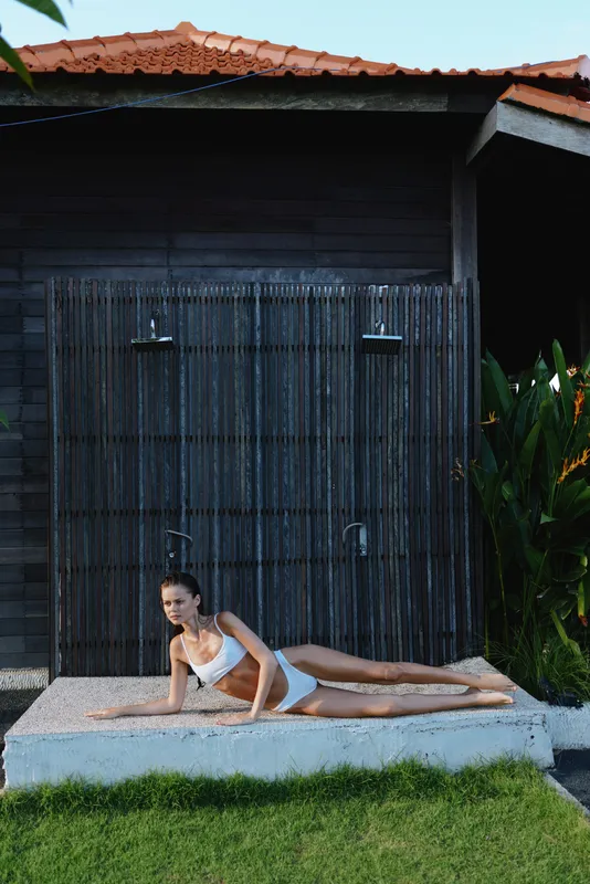 Yoga pose performed by a woman in a white athletic top on a mat outdoors beside a wooden shed, sunlight on grass and water, highlighting lifestyle, fitness, and serenity.