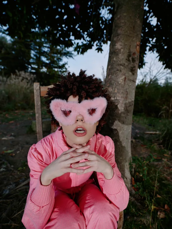 Child in pink outfit with a pink eye mask sits by a tree outdoors, hands folded under the chin, a dreamy costume scene blending nature and imagination.