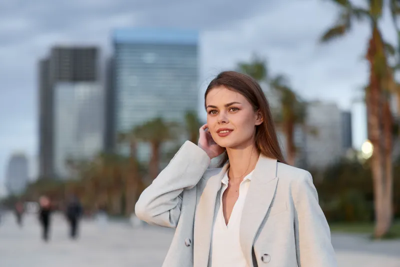 Young woman in light gray coat confidently posing outdoors in urban environment with palm trees and skyscrapers, stylish business fashion concept