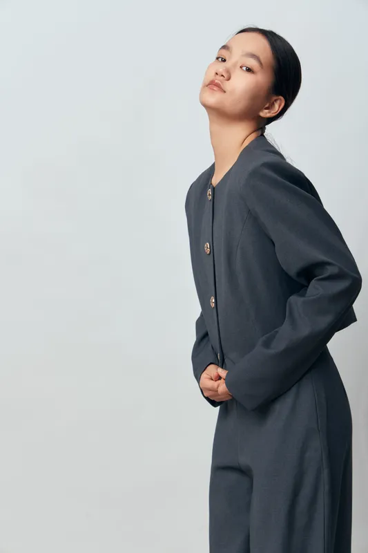 Young woman in gray outfit posing with hands clasped behind back against plain light background for fashion, style and portrait photography studio concept.