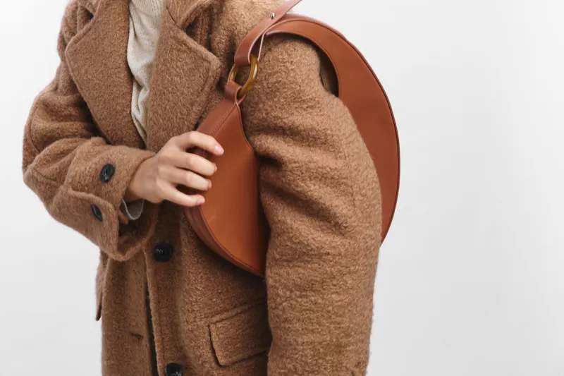 Asian woman wearing a warm brown coat with a round leather bag stands against a plain background, highlighting fashion, texture, and poise in a modern lifestyle scene.