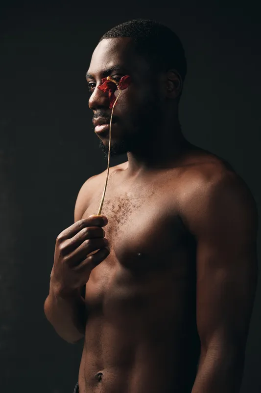 Black man with dark skin holding dried flower near face in creative visual designer style, artistic portrait with moody lighting and emotional expression on dark background.