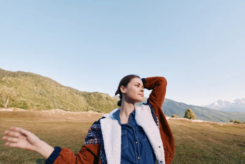 Woman portrait outdoors in mountains landscape and open field, wearing warm jacket and denim shirt, enjoying nature and meadow with distant peaks under clear blue sky.