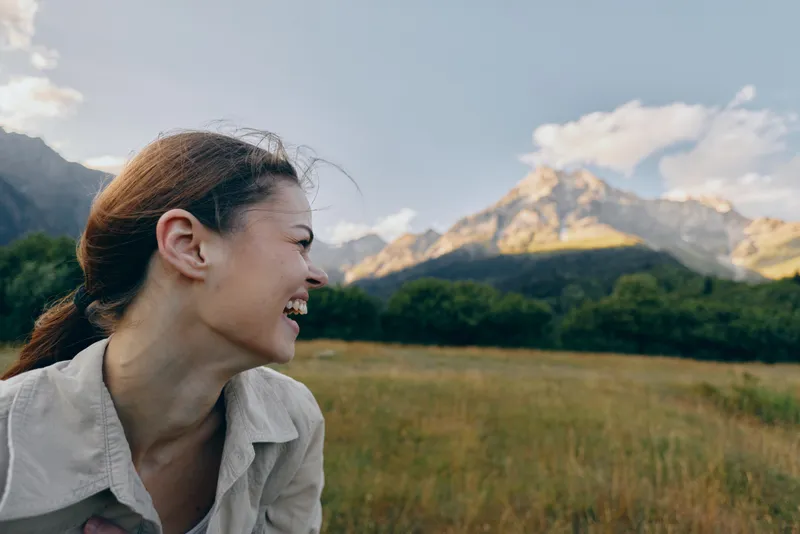 A smiling woman with tied hair enjoys a sunny day outdoors in a grassy field with mountains and blue sky in the background. Lifestyle concept with nature and happiness.