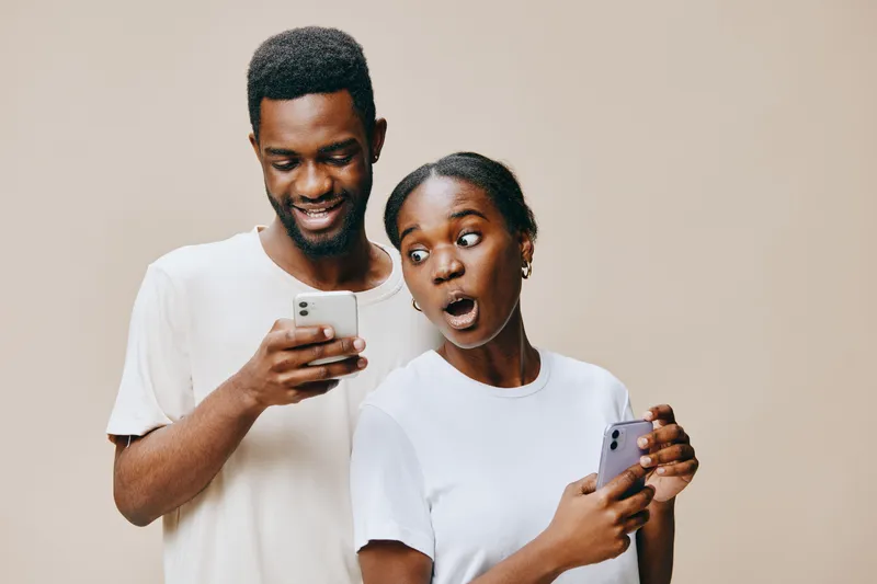 Surprised young couple looking at smartphones with excitement, showcasing joyful emotions in neutral background, perfect for social media and digital communication themes