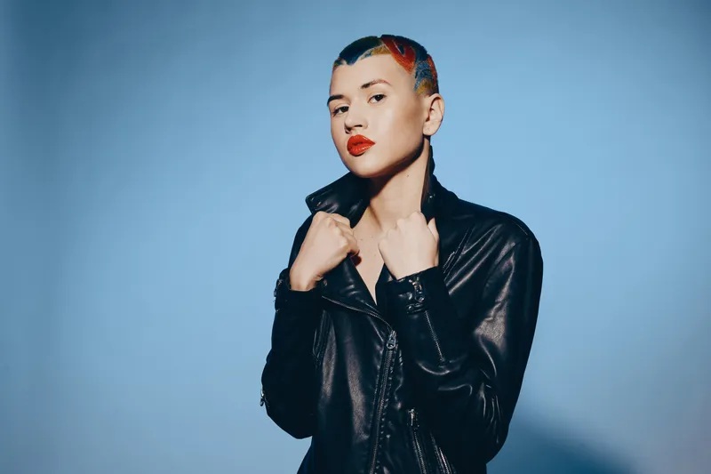 woman with colorful hair wearing black leather jacket stands confidently against blue background with bold makeup and modern edgy style