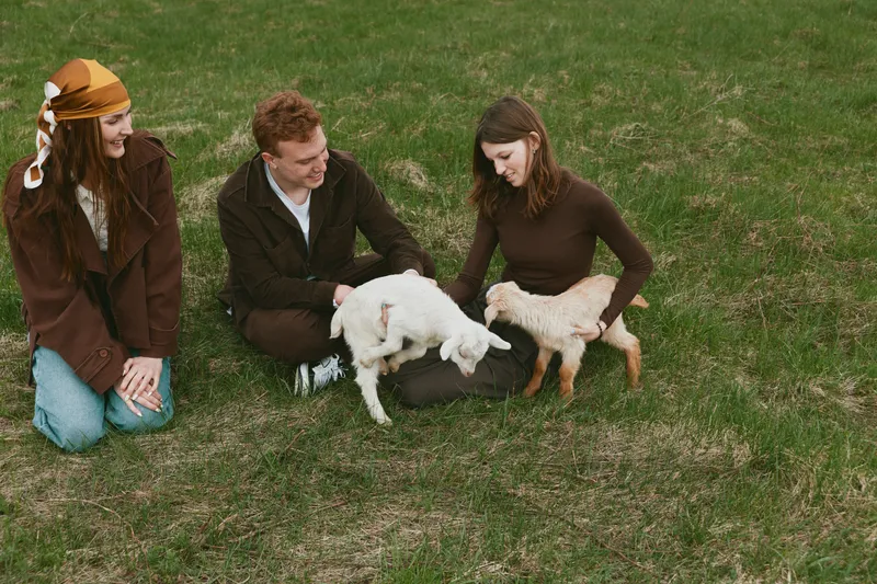 A casual outdoor scene shows three adults on a grassy field interacting with two young goats, pets and companionship. A candid lifestyle moment highlighting bonding, care, and rural simplicity.