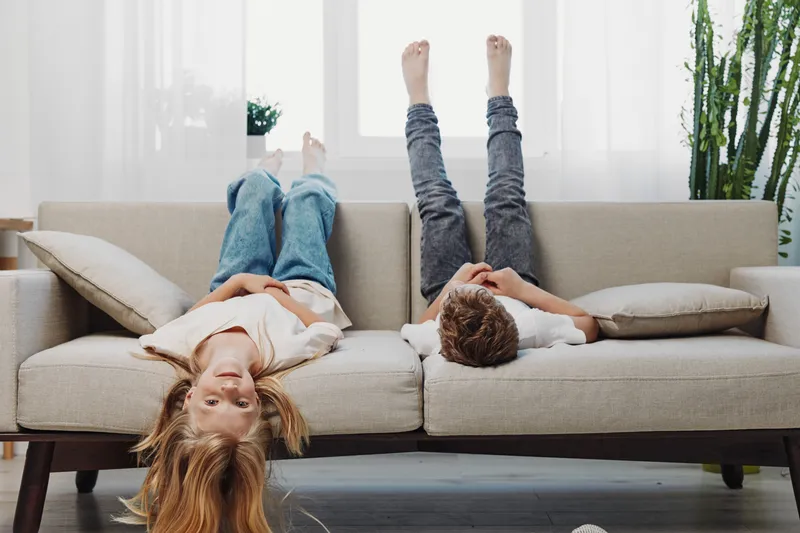 Two children lying upside down on a beige sofa with legs up, relaxing in a bright living room with natural light and green plants in the background, capturing playful home moments.