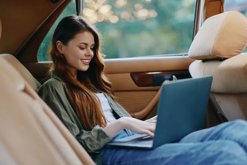 Young woman working on a laptop in a car, wearing casual clothing, smiling with a content expression, lush green background, warm colors, and a relaxed atmosphere.