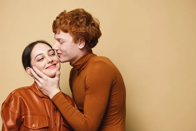 Couple, love and affection with man kissing woman cheek in studio. Young people, romance and happiness with casual clothes and warm emotion on beige background.