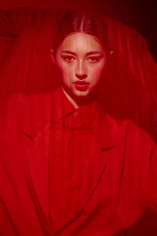 Fashion model in red suit and veil poses with intense expression against dark red background. Creative makeup enhances moody bold beauty concept with artistic lighting.
