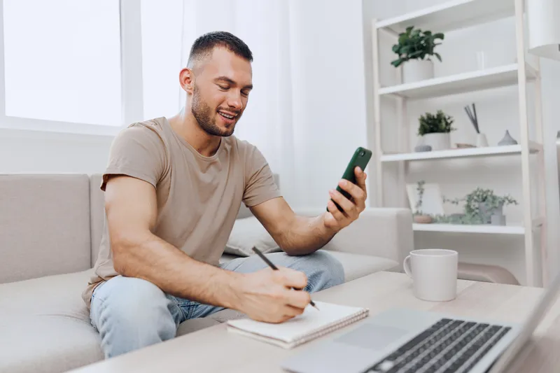 Smiling man sitting on the couch, using smartphone while taking notes. Bright and cozy living room setup, showcasing modern lifestyle trends of 2025.