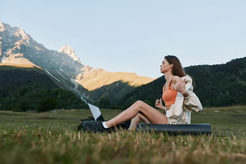 A young woman sits on a grassy field with a laptop, wearing an orange top and light jacket, enjoying outdoor work in a scenic mountain landscape.