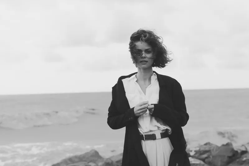 Woman on a windswept beach wearing a tailored coat and white shirt, adjusting her belt as waves roll in, monochrome photography conveying mood and elegance.