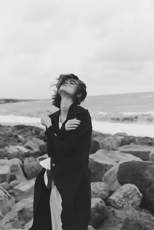 Monochrome beach scene with a woman in a long coat on a windswept shoreline, gazing upward, rugged rocks and ocean in the background, moody weather, contemplative outdoor fashion moment