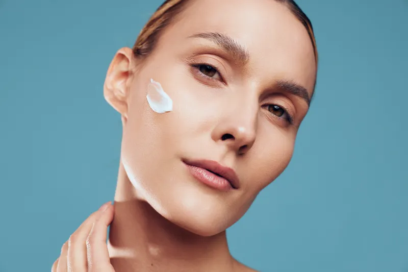 Closeup of woman applying cream on cheek, smooth skin texture, natural makeup, soft lighting, blue background, skincare concept, beauty routine, healthy facial care